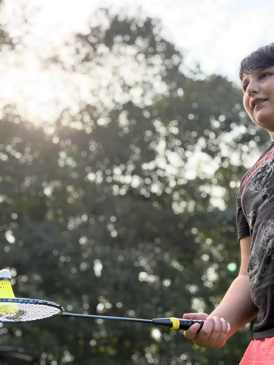 A Scout holding a badminton racket with a shuttlecock resting on the strings, lining up a serve with sunlit trees behind him.