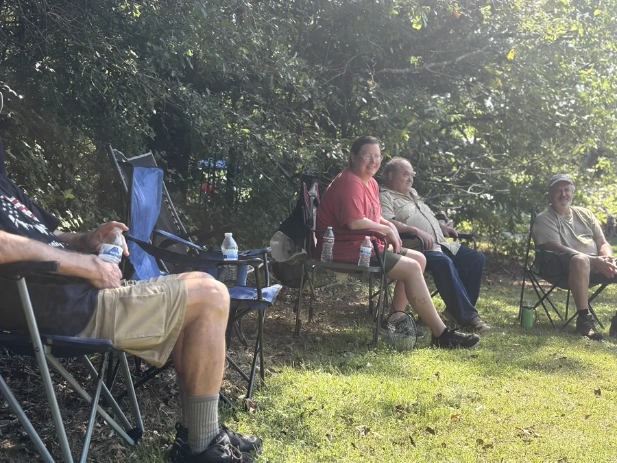 Four adult leaders sitting in a row of camp chairs along a treeline in the afternoon sun, with a badminton racket lying in the grass.