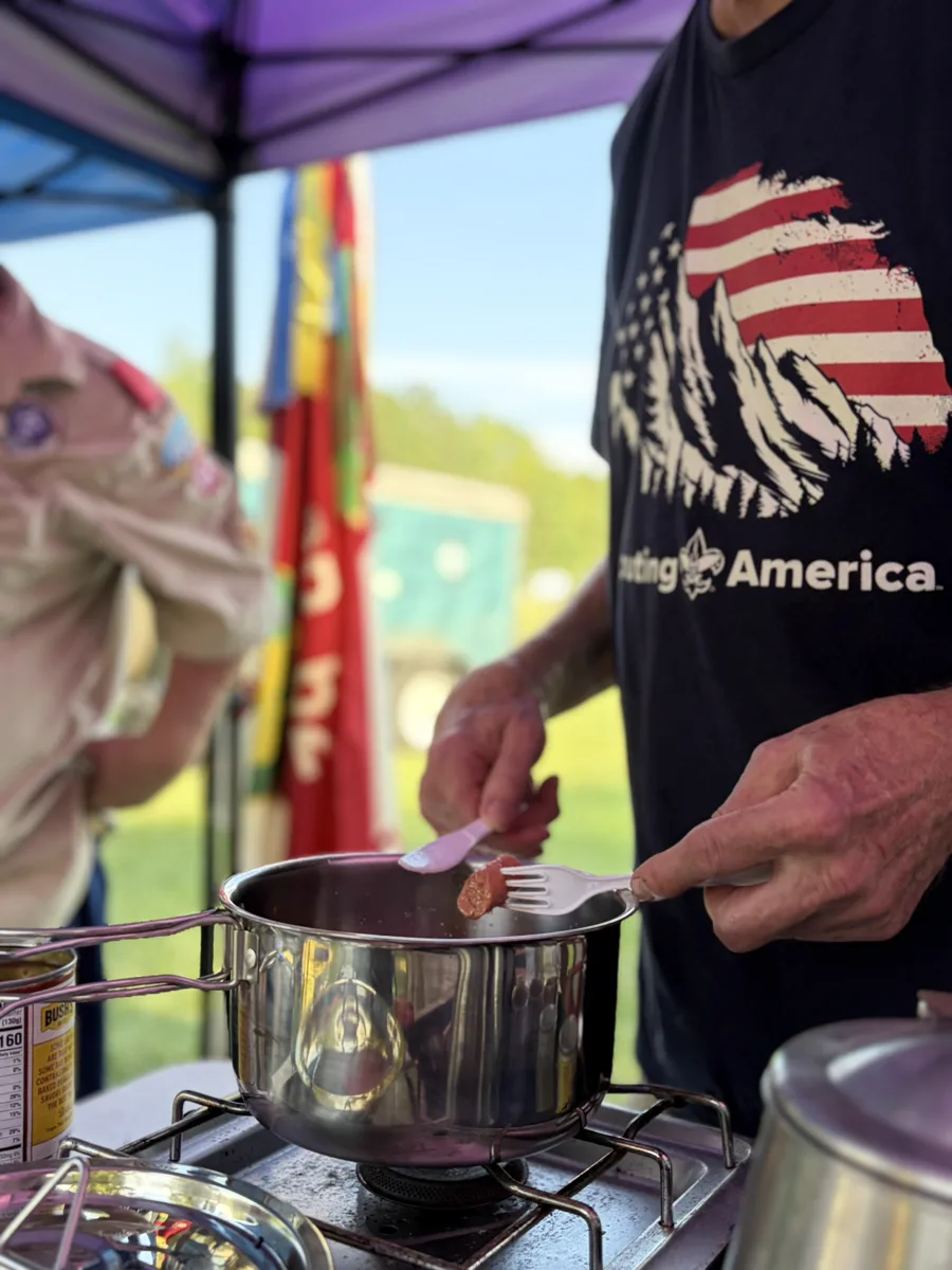 A leader in a Scouting America T-shirt lifting a piece of meat on a slotted spoon out of a pot on a camp stove, under a canopy with troop flags behind him.