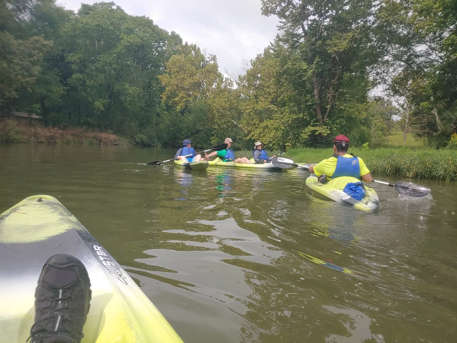 Scouts and leaders paddling kayaks down a tree-lined creek on an overcast summer morning.