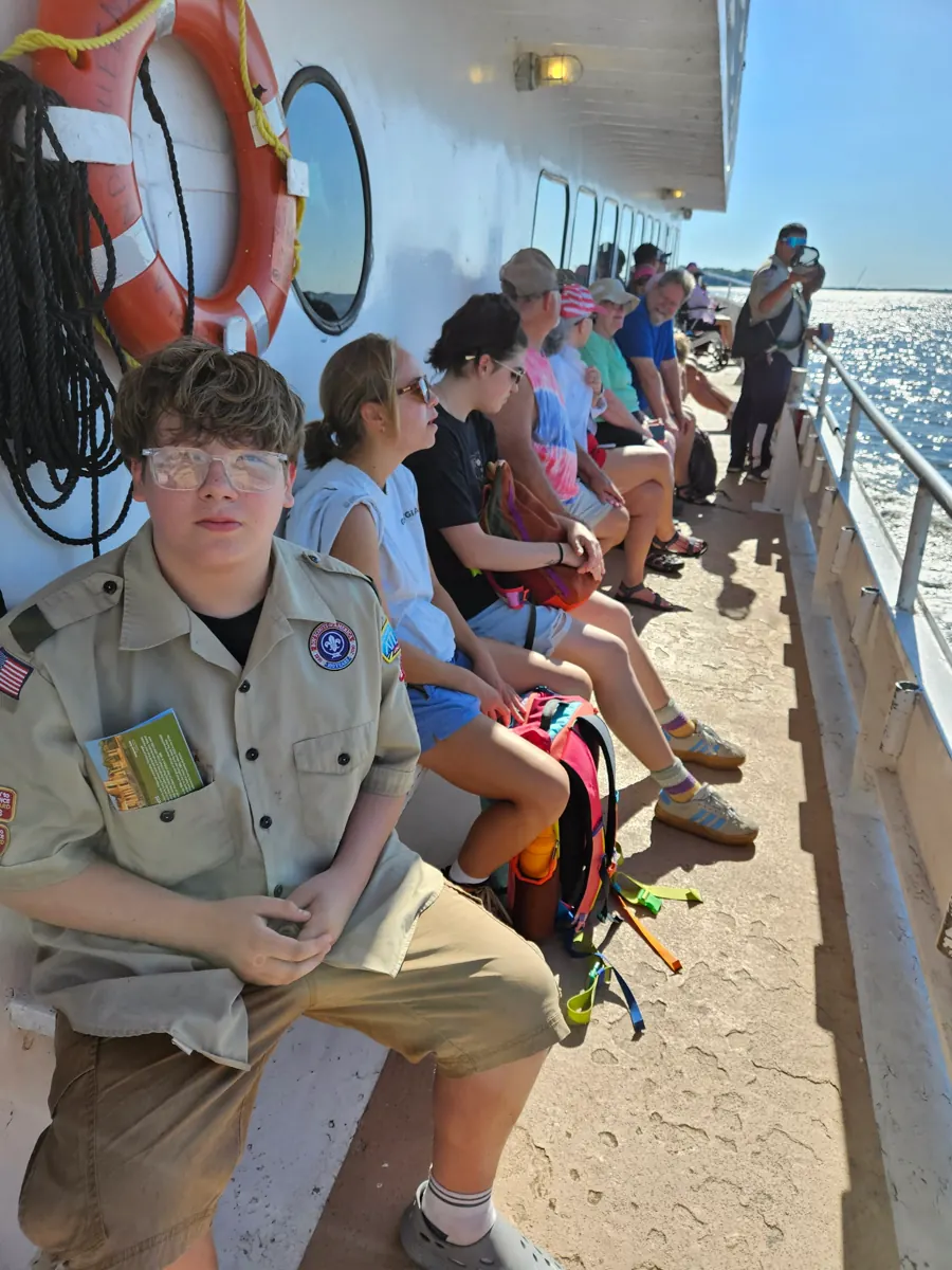 A Scout in uniform sitting on the open deck of a ferry, other passengers along the rail and bright water beyond.