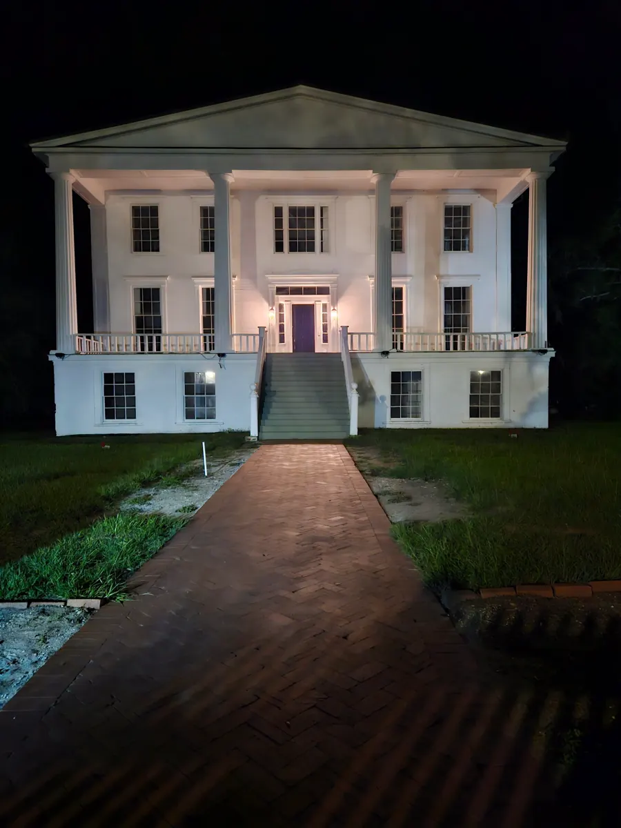 A large white house with a columned portico lit up at night at the end of a brick path.
