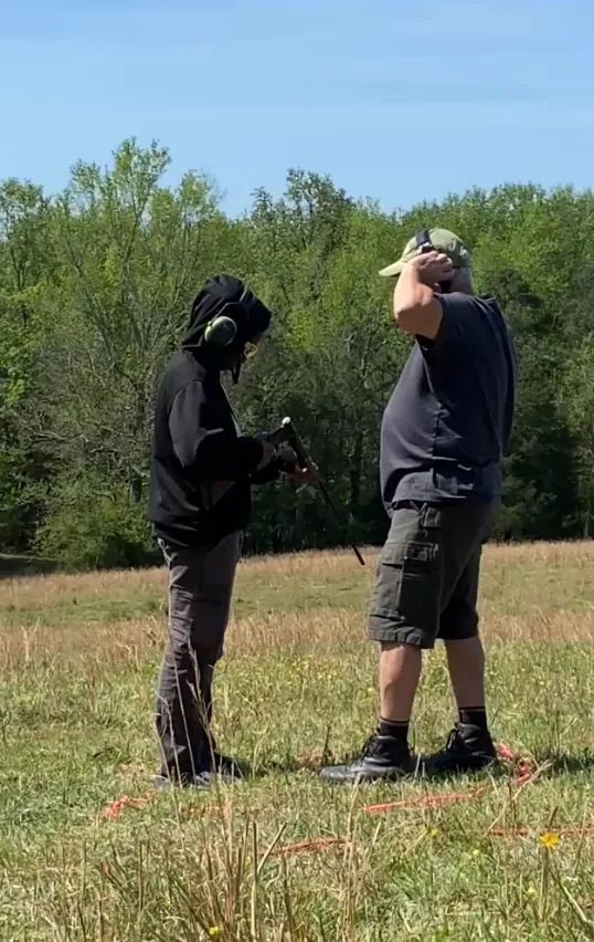A Scout in ear protection being coached on the firing line by an adult range instructor in an open field.