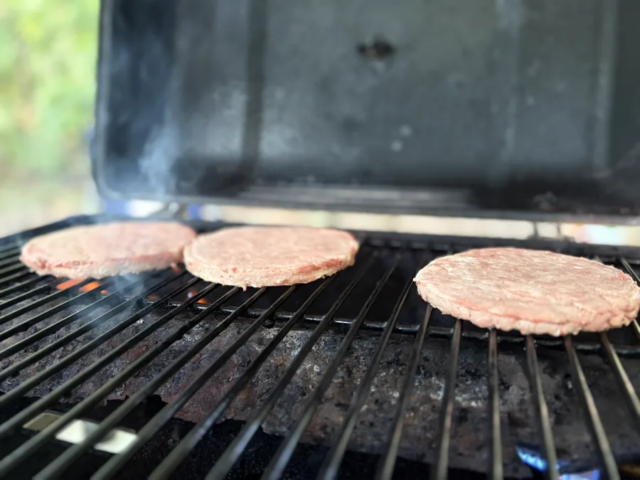 Burger patties cooking on the grate of a camp grill with the lid propped open and woods behind.