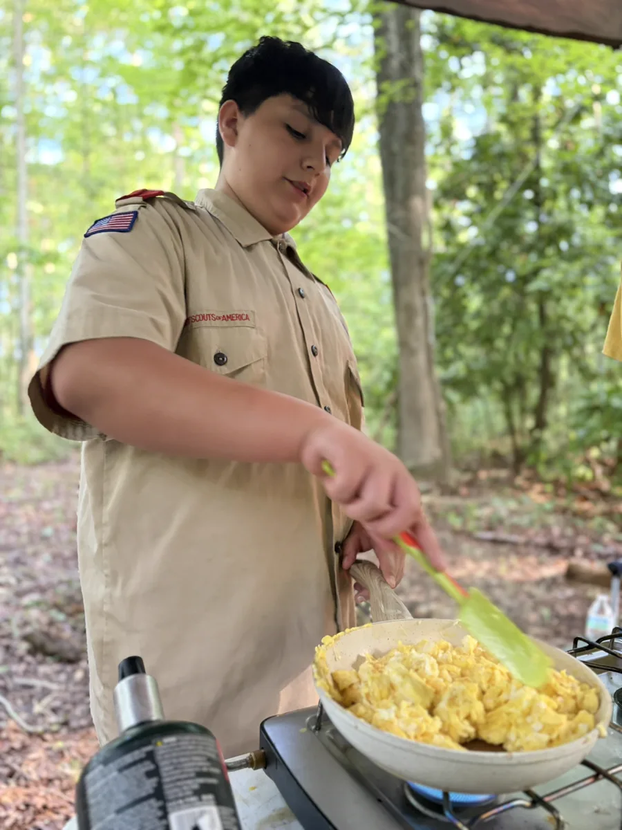 A Scout in uniform stirring a pan of scrambled eggs on a camp stove under a shelter in the woods.