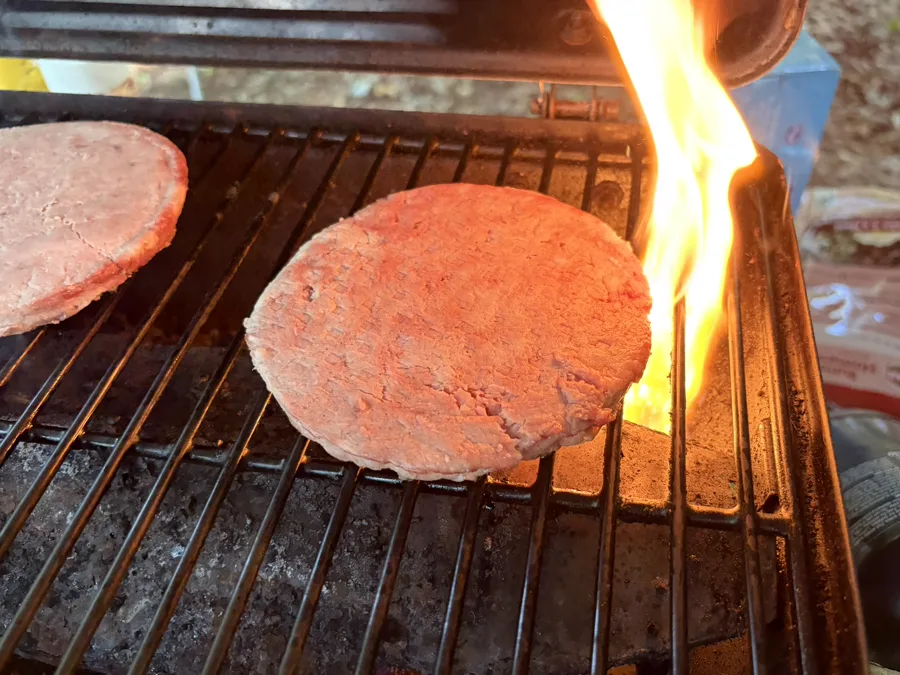A burger patty on a grill grate with a tall jet of flame rising beside it during a flare-up.