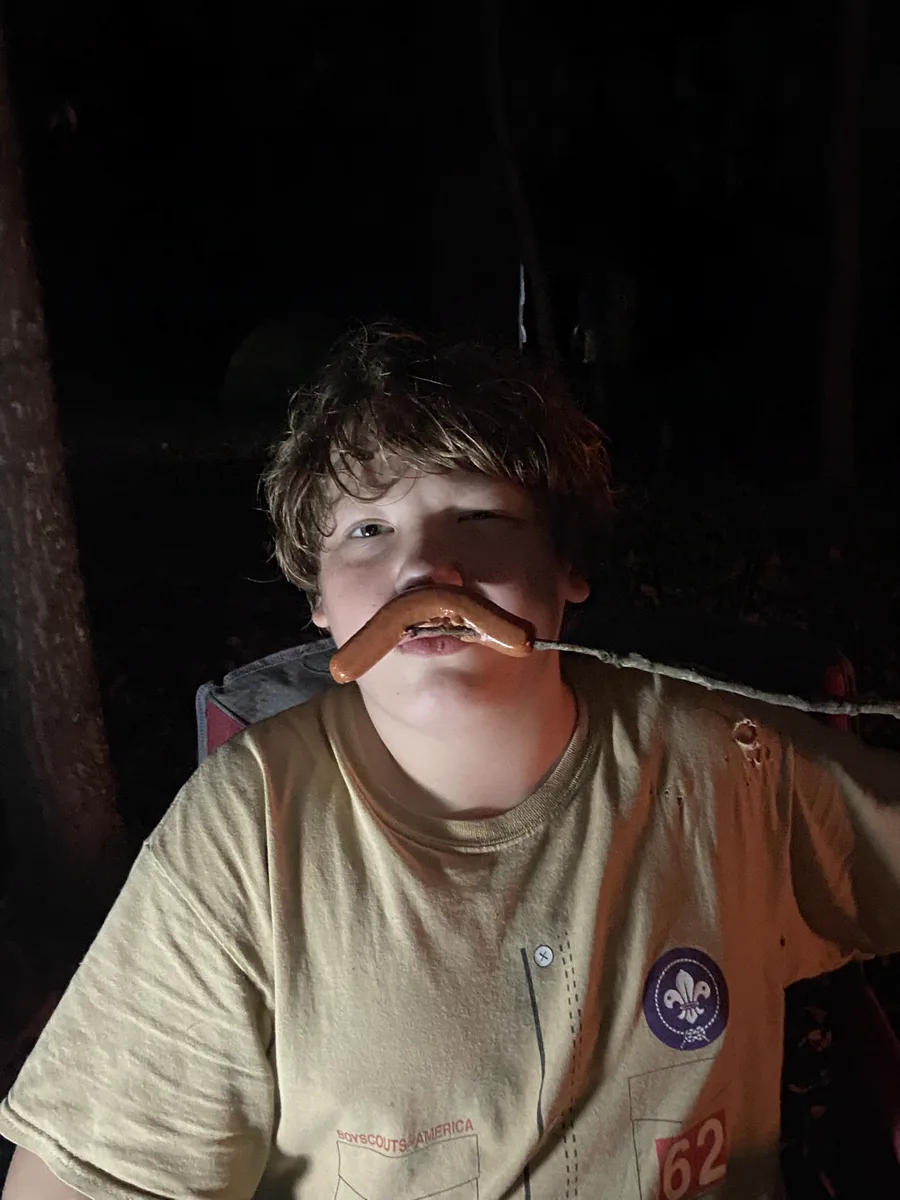 A Scout at a night-time campfire holding a roasted hot dog on a stick up under his nose like a moustache, wearing a Boy Scouts of America T-shirt with a troop 62 numeral.