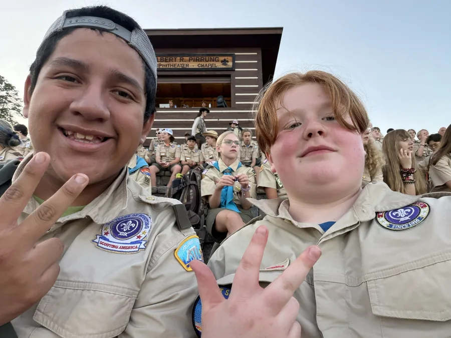Two Scouts in uniform grinning at the camera on the amphitheater steps as Scouts from other units fill the benches behind them.