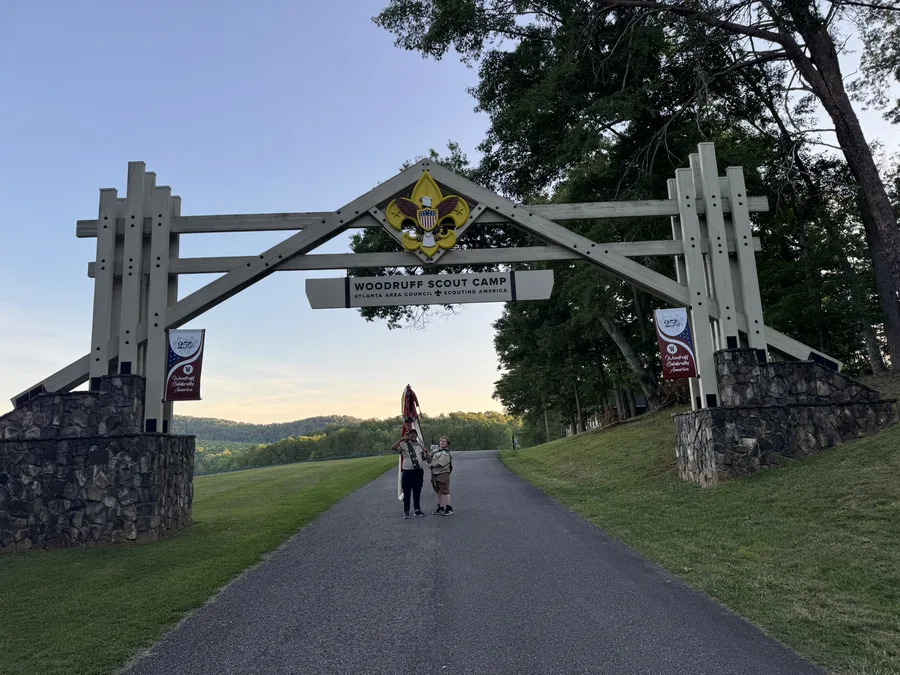 Two Scouts standing under the timber entrance arch of Woodruff Scout Camp at sunrise, one holding the troop flag, with wooded ridges behind them.